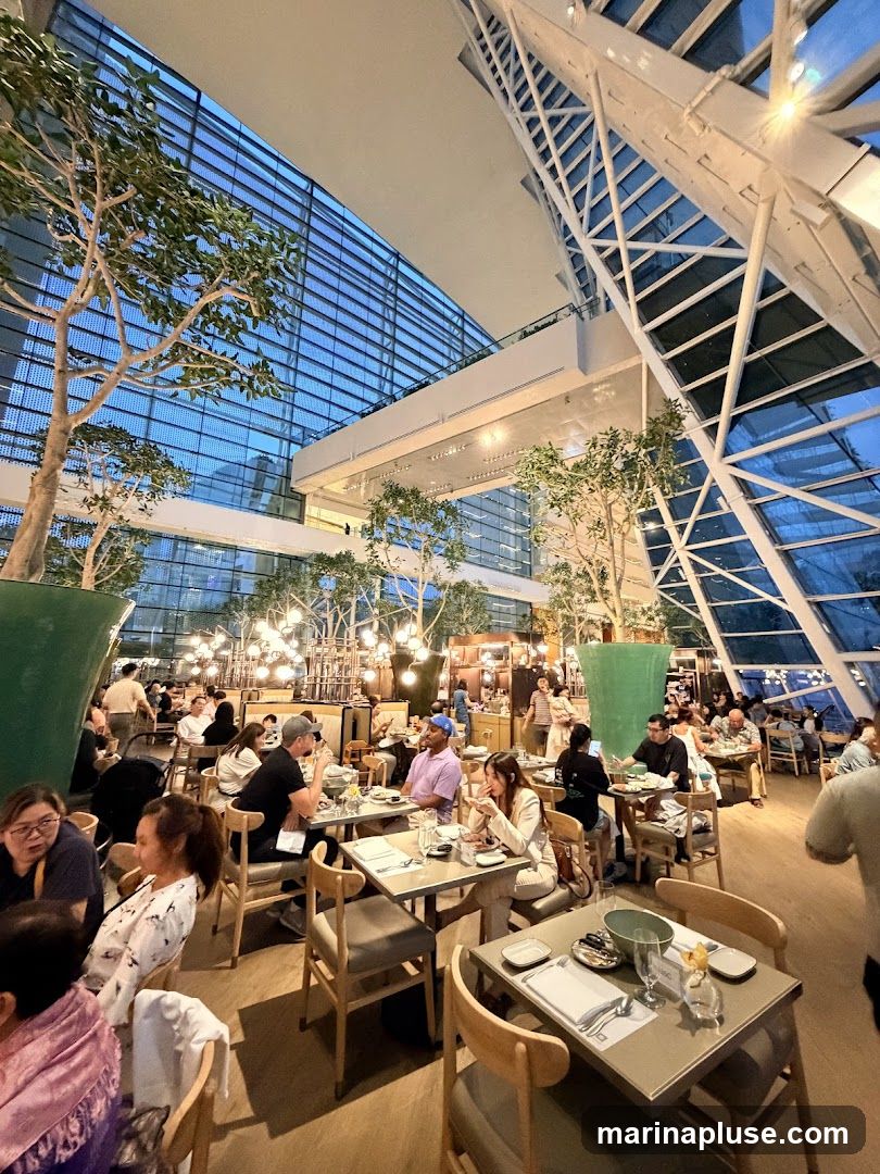 A spacious restaurant interior with high ceilings and large windows, featuring numerous tables occupied by diners. The decor includes indoor trees and ambient lighting.