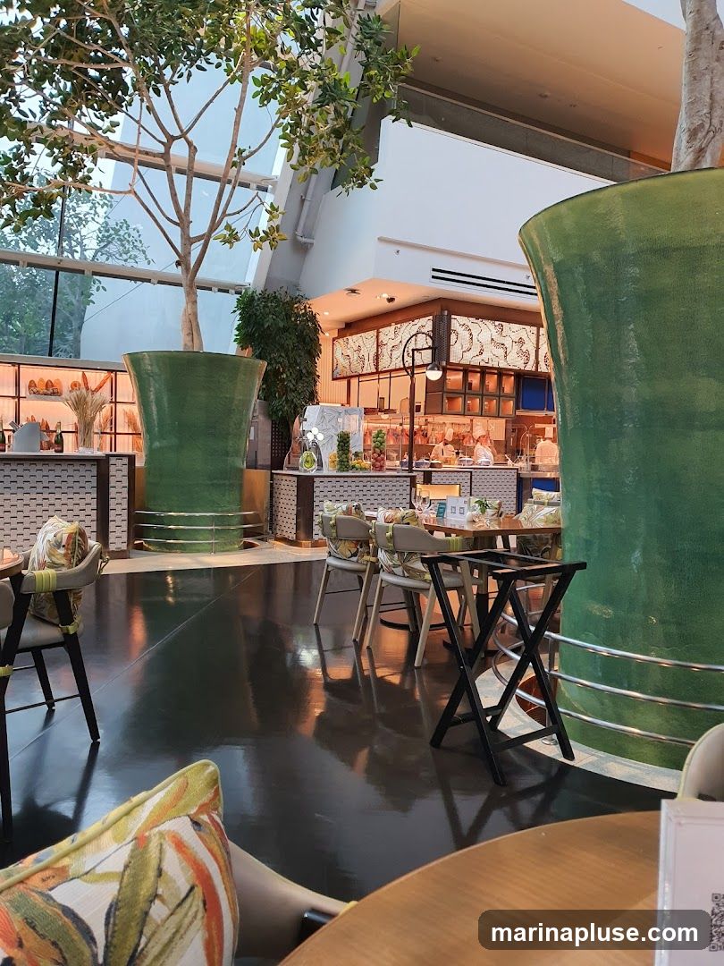 The image shows the interior of a restaurant with a modern and airy design. Large, vibrant green planters with trees are placed strategically throughout the space. The flooring is a dark, reflective surface, creating a sense of depth. Tables and chairs with tropical-patterned upholstery are set up, suggesting a comfortable dining area. In the background, a food service counter is visible with staff members preparing dishes. The overall ambiance is sophisticated and inviting, with natural light filtering in from a large window or skylight.