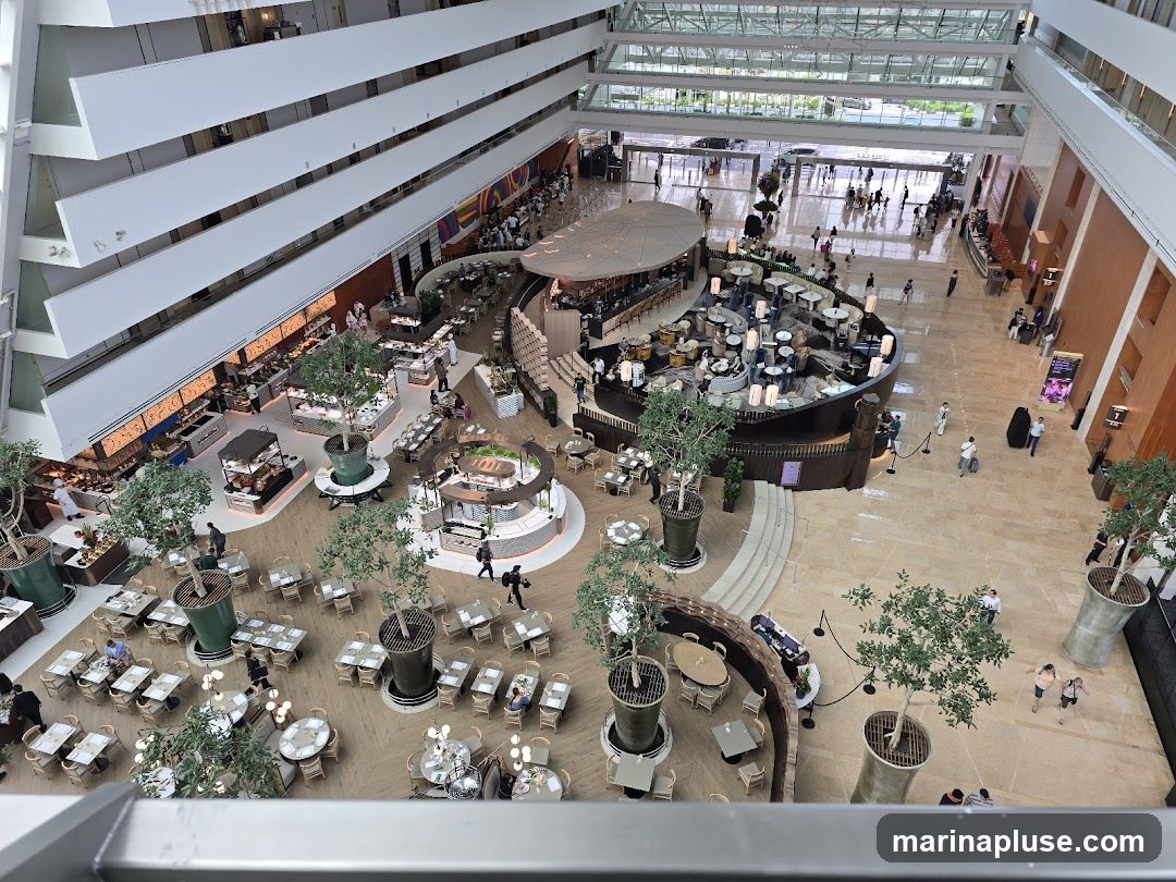An aerial view of a large, multi-level hotel atrium with a central dining area. The dining area features a circular bar and seating arrangements, surrounded by buffet stations and tables with chairs. Potted trees are strategically placed throughout the space. People are seen walking and dining. The atrium has a modern design with white balconies overlooking the central area.