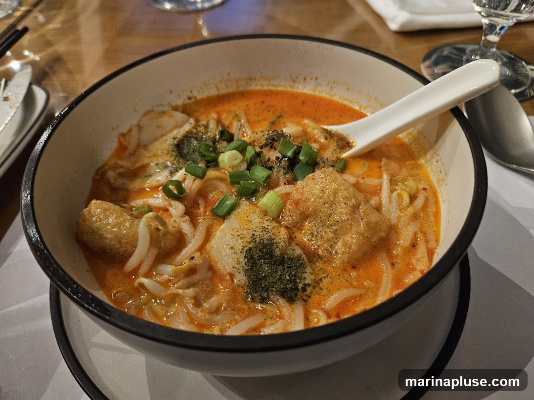 A close-up shot of a bowl of laksa noodle soup, featuring a rich orange broth, noodles, bean sprouts, fried tofu puffs, and chopped green onions. A white spoon rests in the bowl. The dish is served on a table with cutlery and glassware visible in the background.