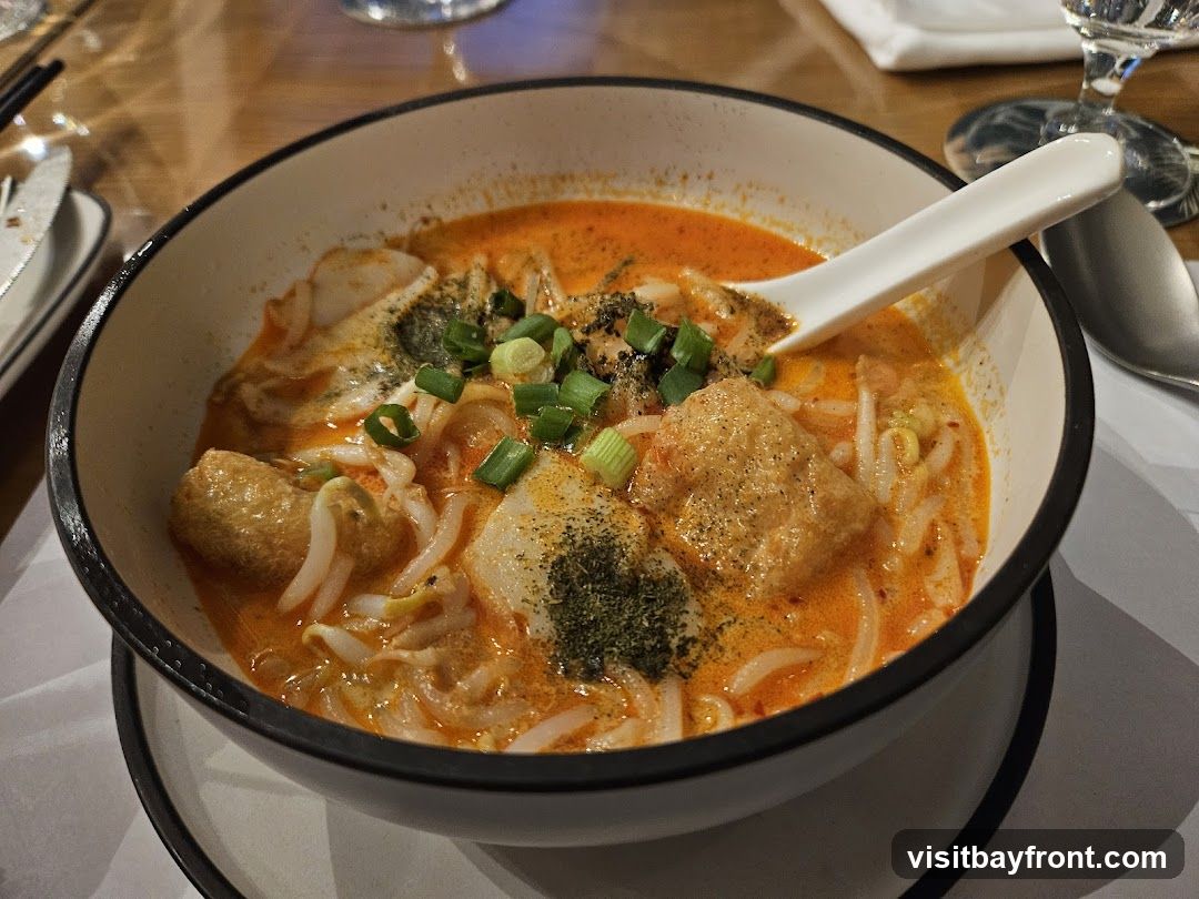 A close-up shot of a bowl of laksa noodle soup, featuring a rich orange broth, noodles, bean sprouts, fried tofu puffs, and chopped green onions. A white spoon rests in the bowl. The dish is served on a table with cutlery and glassware visible in the background.