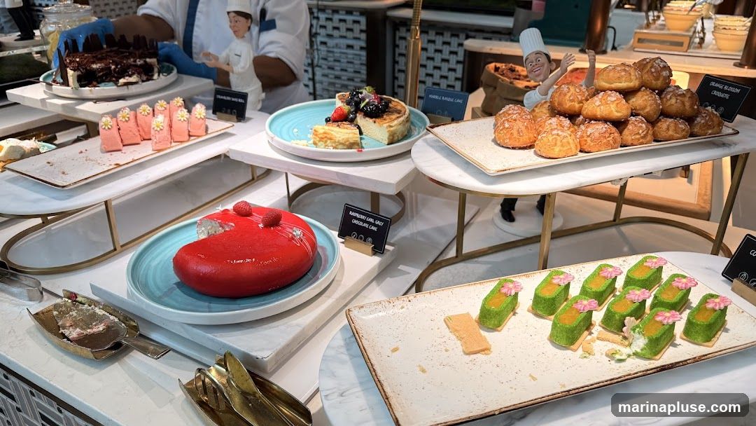 A variety of desserts are displayed on a buffet table, including cakes, pastries, and small individual treats. Some desserts have labels indicating their names. A chef figurine is placed near a pile of cream puffs.