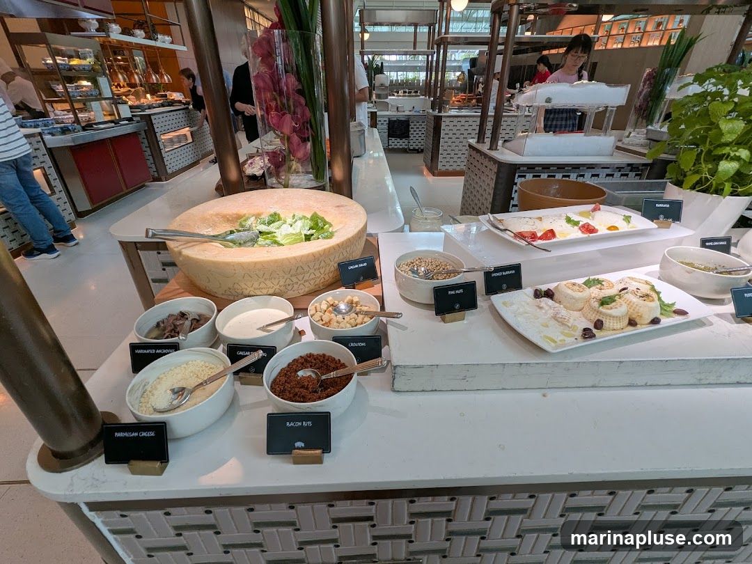 A buffet salad bar with various toppings and ingredients. In the foreground, a large wheel of cheese is hollowed out and filled with salad. Surrounding it are bowls of marinated anchovies, Caesar dressing, croutons, parmesan cheese, and bacon bits. Further back, there are platters of food, including what appears to be burrata and other cheeses, along with pine nuts and a green sauce. In the background, people are serving themselves from other buffet stations.