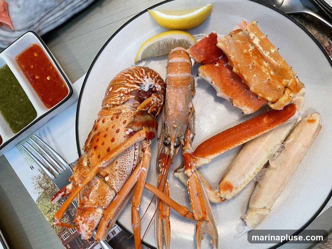A plate of assorted seafood including a langoustine, crab legs, and what appears to be a small lobster. Two dipping sauces, one red and one green, are served in a small rectangular dish. Lemon wedges are also on the plate. A fork rests on a placemat with an image of buildings.