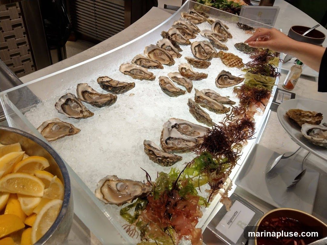 A close-up view of a buffet display featuring a large selection of fresh oysters on ice. The oysters are arranged in rows, some open and some closed. Garnishes of seaweed and lemon wedges are also visible. A hand is reaching for an oyster, and a small sign labeled "mignonette" is present. In the foreground, a bowl of sliced lemons is prominent.