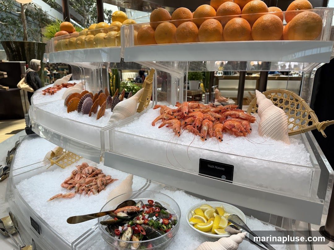 A buffet display featuring various seafood items on ice. In the foreground, a bowl of mussels with chopped peppers and herbs is visible, alongside a plate of lemon wedges. Above, trays of cooked prawns and scallops are arranged. The top tier displays a selection of oranges and lemons.