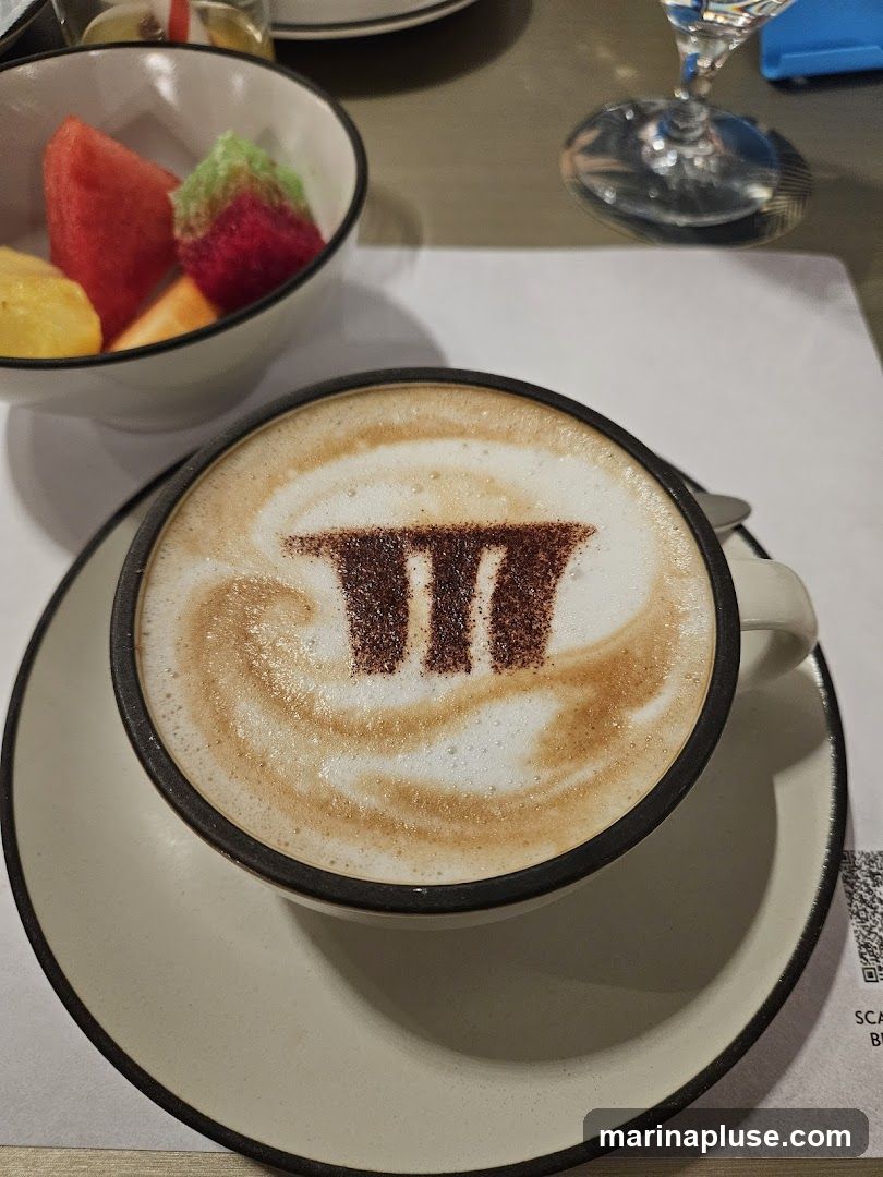 A close-up shot of a cappuccino with latte art featuring a logo made of cocoa powder. The coffee is served in a white mug with a dark rim, placed on a matching saucer. In the background, a white bowl contains a colorful assortment of cut fruits, including watermelon, pineapple, and what appears to be honeydew melon. A wine glass and a table setting are also visible. A QR code is printed on the paper placemat under the saucer.