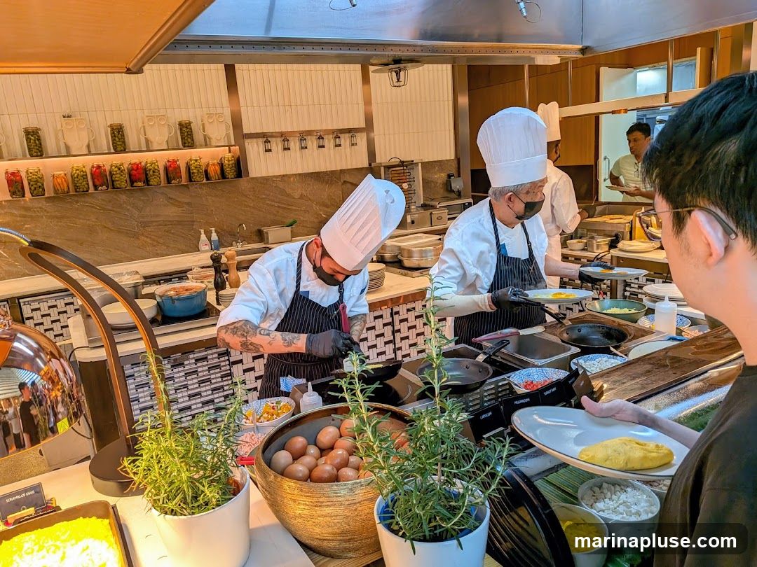 Two chefs in white hats and aprons are preparing food at a buffet station. One chef is cooking an omelet, while the other is attending to a pan. There are various ingredients and dishes laid out on the counter, including eggs, chopped vegetables, and a pot of food. A customer is holding a plate, waiting to be served. The background shows shelves stocked with jars of preserved food and kitchen equipment.