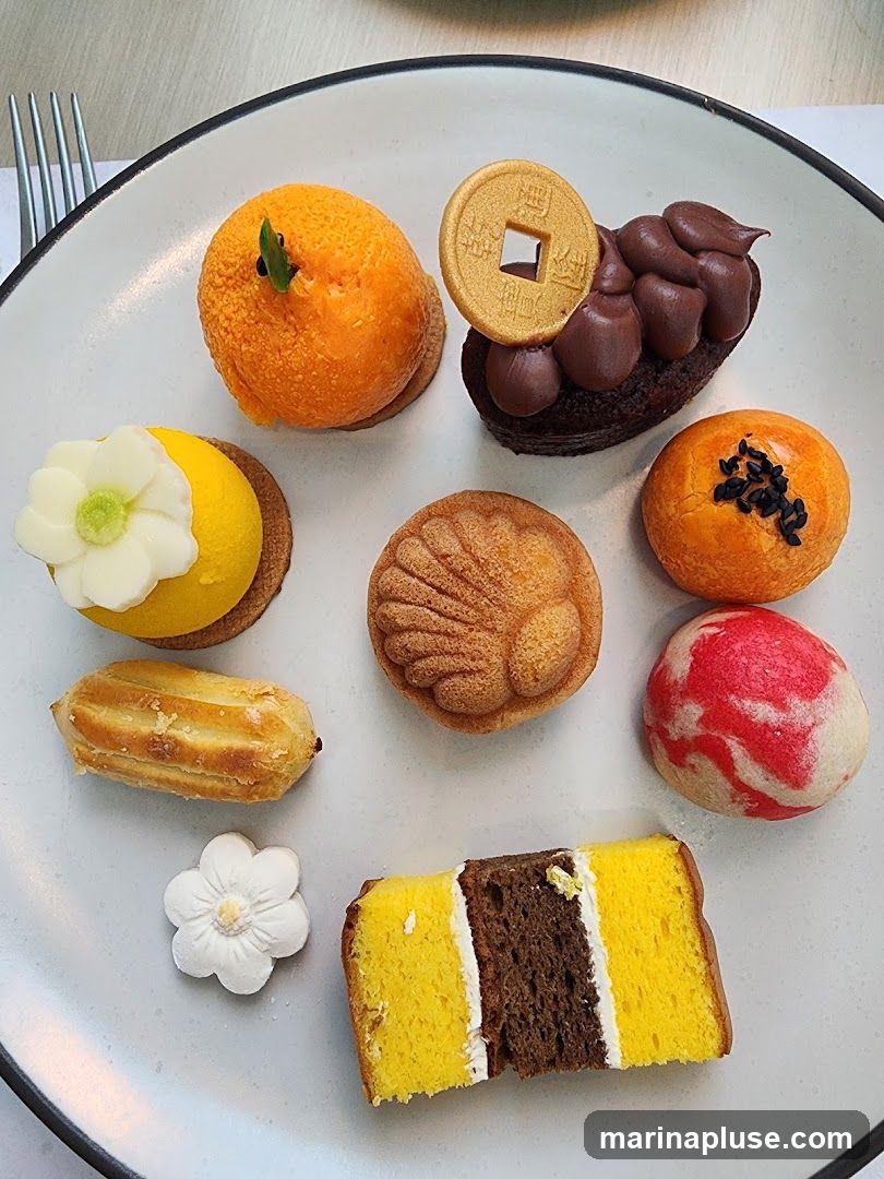 A plate of assorted small desserts, including a cake shaped like an orange, a chocolate pastry with a gold coin decoration, a yellow cake with a white flower, a shell-shaped cake, an eclair, a marble-patterned pastry, a small round pastry with black seeds, and a slice of layered cake with yellow and brown sponge and white cream.