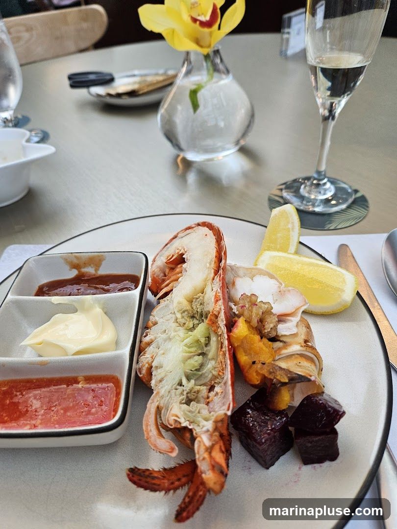 A plate of cooked lobster is served with three small dishes of condiments: a dark red sauce, a creamy white sauce, and a pinkish sauce. There are also lemon wedges and diced beets on the plate. The setting appears to be a restaurant, with a glass of white wine and a small vase with a yellow flower visible in the background.