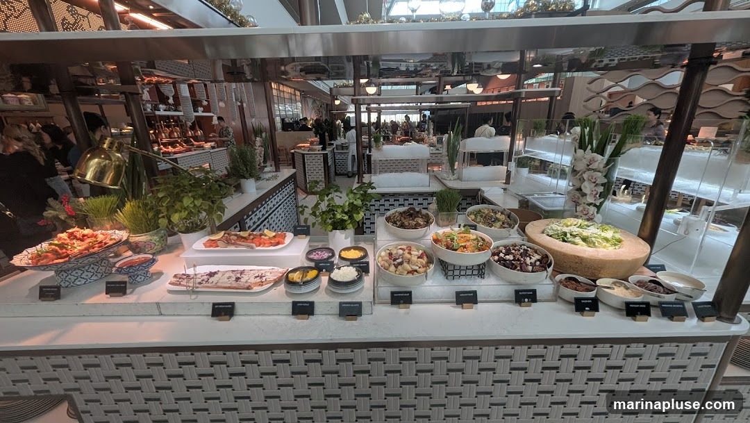 A wide shot of a buffet with various dishes displayed on a counter. In the foreground, there are platters of food, including what appears to be seafood and salads. Several bowls contain different types of salads and side dishes. A large wheel of cheese with a salad inside is prominently featured. In the background, more food stations and people can be seen.
