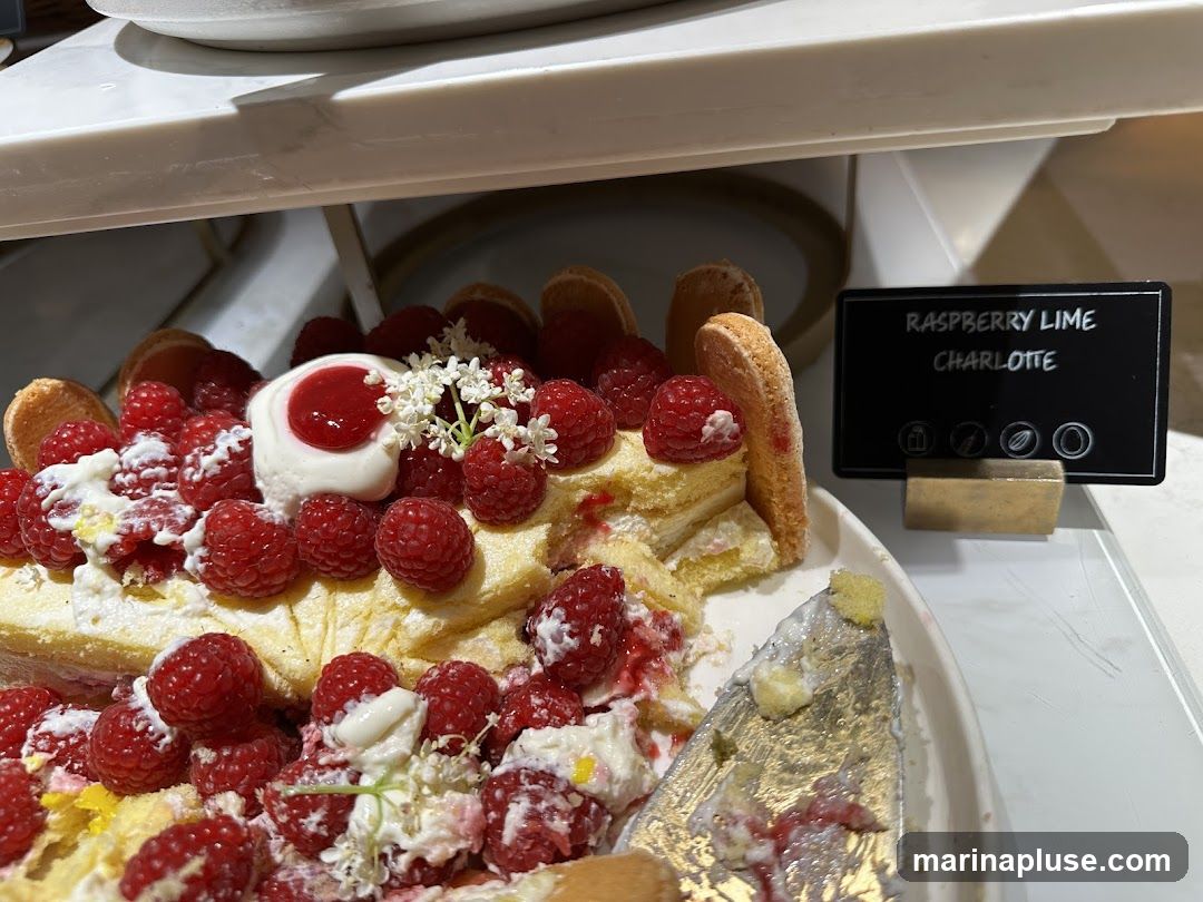 A close-up view of a Raspberry Lime Charlotte dessert on a white platter. The dessert is adorned with fresh raspberries, whipped cream, and delicate white flowers. A small black sign with white text identifies it as "RASPBERRY LIME CHARLOTTE" and includes icons representing ingredients or dietary information. A serving utensil with remnants of the dessert is visible on the right side of the platter.