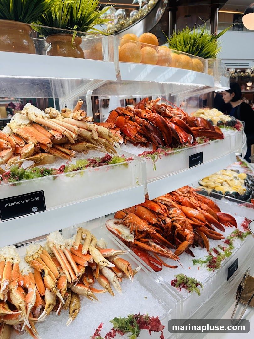 A close-up view of a seafood buffet display featuring an abundance of fresh seafood on ice. In the foreground, piles of snow crab legs are arranged, their orange and white shells contrasting with the clear ice. To the right, a generous portion of cooked lobsters, their vibrant red shells glistening, are displayed. In the background, other seafood items and decorative elements like oranges and greenery are visible. A person is partially visible in the background, suggesting a dining environment.