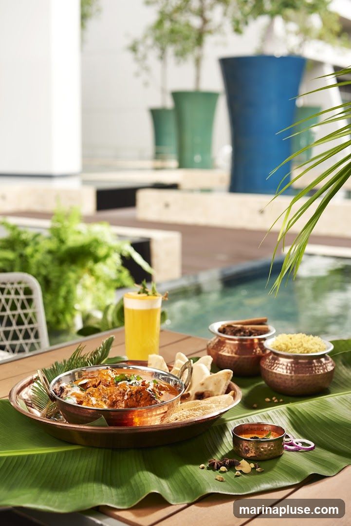 A close-up shot of an Indian food platter, featuring a creamy curry dish in a copper bowl, served with naan bread and a side of rice. A yellow drink with a garnish sits in the background, along with other copper bowls containing spices and rice. The setting appears to be outdoors, possibly a patio or poolside area, with lush greenery and decorative planters visible.