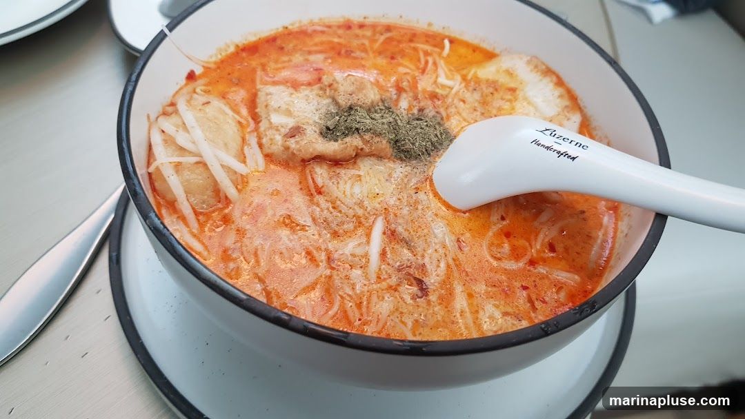 A close-up shot of a bowl of laksa noodle soup. The soup is a rich orange color, with noodles, bean sprouts, and pieces of tofu visible. A white spoon with the text "Luzerne Handcrafted" is resting in the bowl. The dish is garnished with dried herbs.
