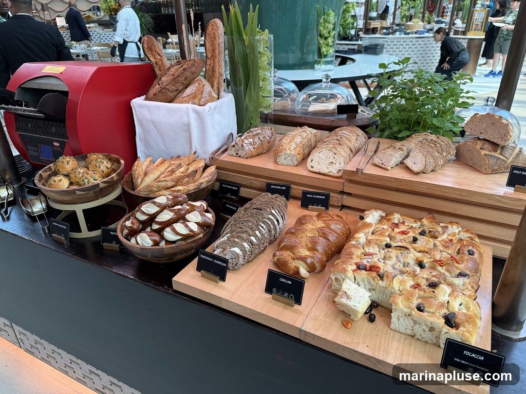 A variety of breads are displayed on a buffet table, including baguettes, pretzels, focaccia, and sliced loaves. A red bread oven is visible on the left.