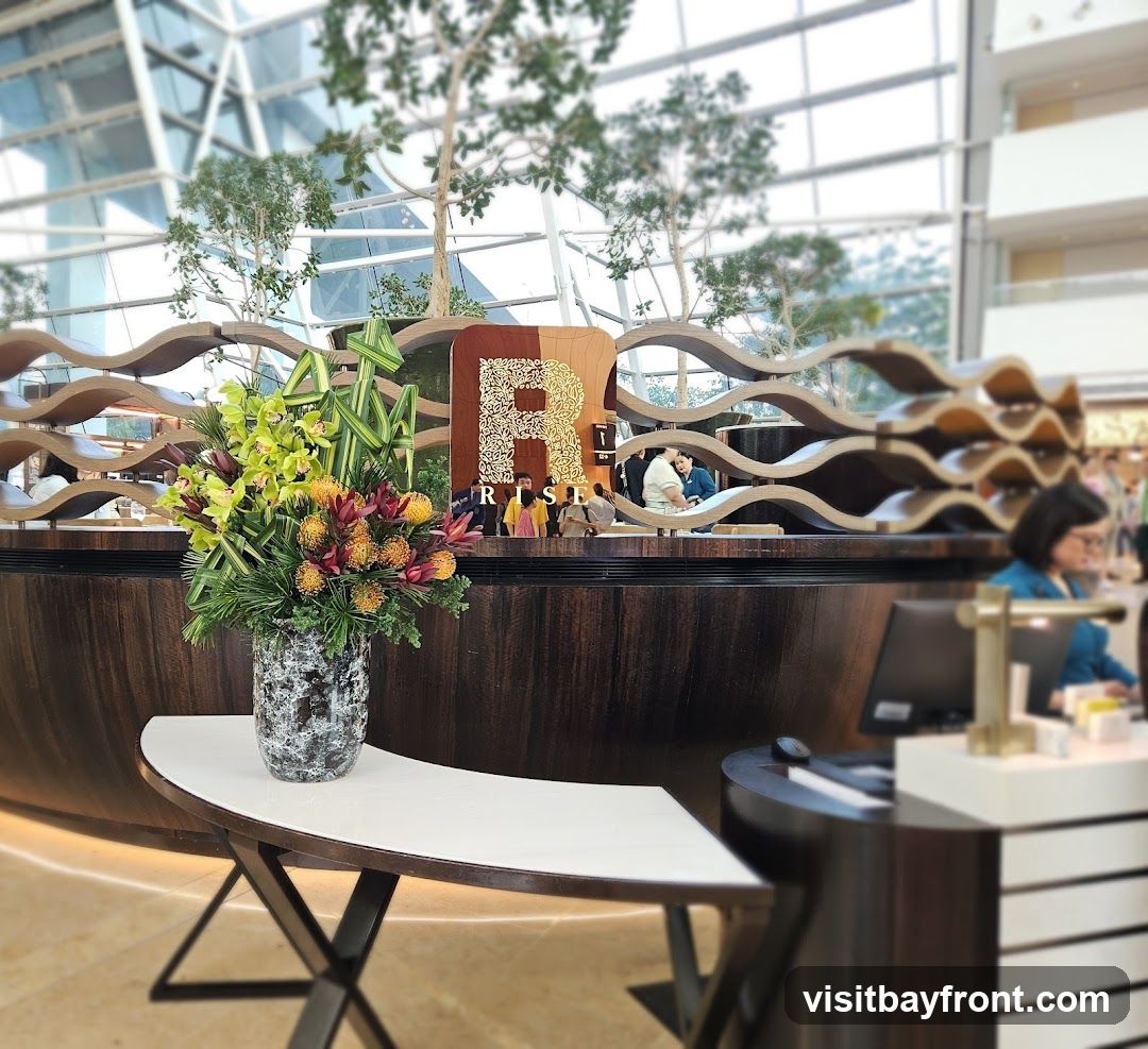 A decorative floral arrangement sits on a white table in front of a wooden counter. Behind the counter, a large sign with the letter 'R' and the word 'RISE' is visible. The background shows a modern interior with trees and a glass ceiling.