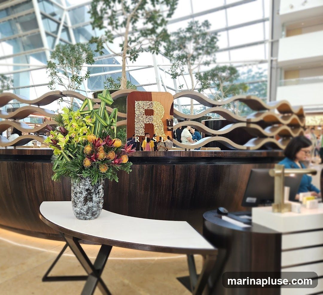 A decorative floral arrangement sits on a white table in front of a wooden counter. Behind the counter, a large sign with the letter 'R' and the word 'RISE' is visible. The background shows a modern interior with trees and a glass ceiling.