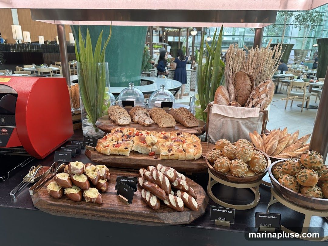 A buffet-style display of various breads and pastries, including loaves, rolls, and pretzels. A red bread slicer is visible on the left. In the background, a restaurant dining area with tables and chairs can be seen.