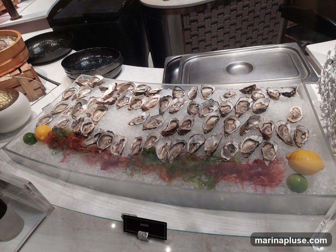 A display of fresh oysters on a bed of crushed ice, garnished with seaweed, lemon, and lime wedges. The oysters are arranged in rows, ready for serving. In the background, there are serving dishes and a metal chafing dish. A small black sign reads "OYSTERS".