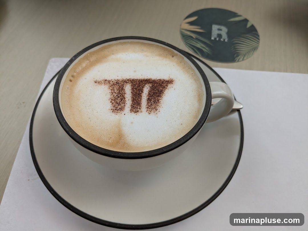 A close-up shot of a cappuccino with the mathematical symbol pi dusted on top with cocoa powder. The coffee is served in a white mug with a black rim, placed on a matching saucer. In the background, a round coaster with a tropical leaf pattern and the letter R is visible.