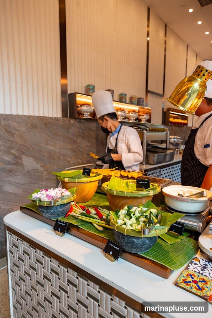 Chefs in uniform prepare food at a buffet station. In the foreground, there are various dishes displayed on banana leaves, including sliced red onion, chilies, lemongrass, and sliced cucumber. In the background, skewers of food are being prepared, and decorative jars are visible on shelves.
