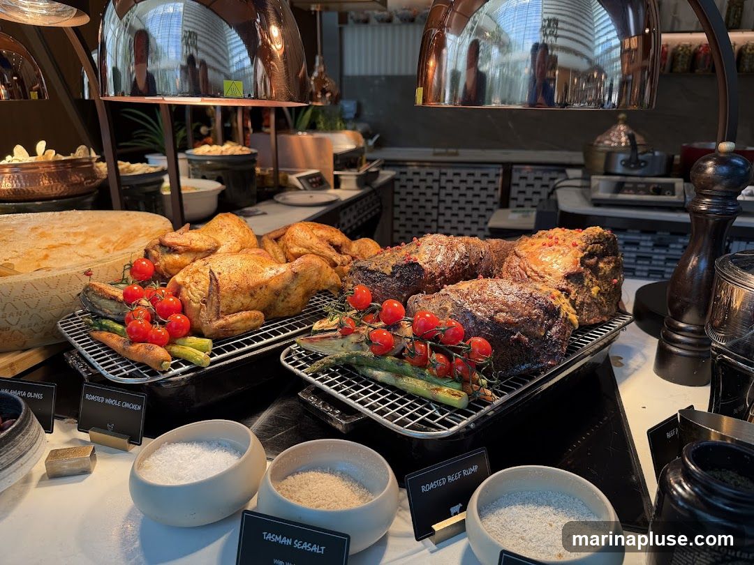 A buffet spread featuring roasted whole chickens and roasted beef rump, garnished with cherry tomatoes and asparagus. Several bowls of salt are placed in front of the meats, labeled 'Tasman Seasalt' and 'Kalamata Olives'. In the background, there are other food items and kitchen equipment.