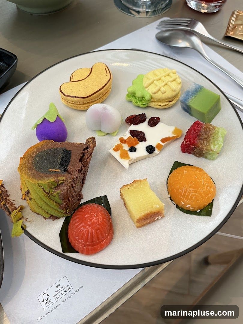 A plate of assorted desserts including macarons shaped like a cashew and a pineapple, a purple eggplant-shaped confection, a peach-shaped confection, white chocolate bark with dried fruit, a slice of green tea cake, a red molded dessert, a slice of yellow cake, and a round orange dessert.