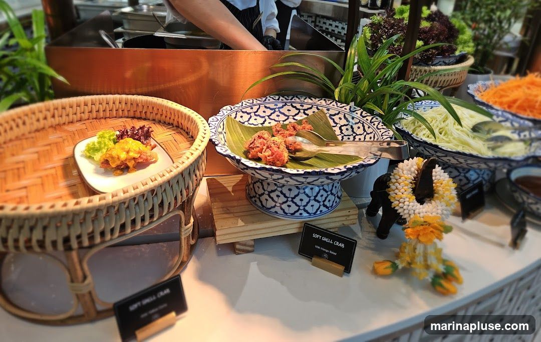 A close-up view of a buffet spread featuring several dishes. In the foreground, a woven basket holds a plate with a piece of food topped with yellow sauce and garnished with lettuce. Next to it, a decorative bowl on a stand contains fried items served on banana leaves, with serving spoons. Further to the right, a bowl of shredded carrots and a bowl of shredded green mango are visible. A decorative elephant figure adorned with a floral garland stands near the mango salad. In the background, a person in a chef's uniform is partially visible, suggesting food preparation. Small black signs with white text identify some of the dishes.