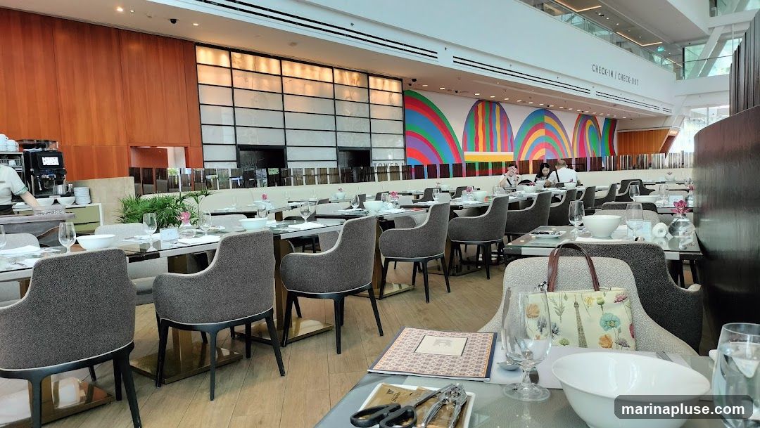 The image shows the interior of a restaurant with tables set for dining. A large, colorful mural featuring abstract arches adorns the back wall. The seating consists of upholstered chairs around glass-topped tables. In the foreground, a table is set with white bowls, glassware, and a menu. A person in a uniform is visible behind a counter with a coffee machine on the left. The overall ambiance is modern and bright, with natural wood paneling on one wall and a view of a modern building structure in the background.