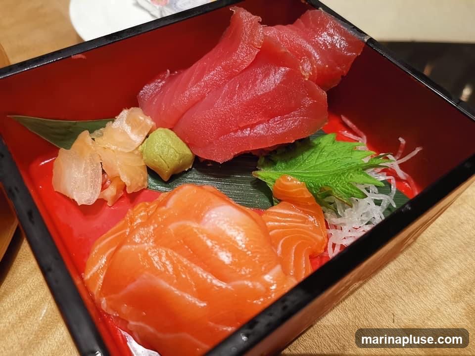 A close-up shot of a sashimi platter featuring slices of salmon and tuna. The platter also includes pickled ginger, wasabi, and garnishes of shiso leaf and shredded daikon radish. The sashimi is arranged in a red and black square dish.