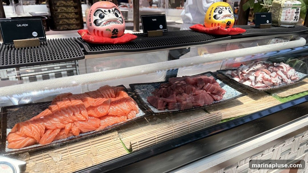 A display case filled with fresh, sliced salmon, diced tuna, and chopped octopus. Decorative Daruma dolls are placed above the display. Small signs indicate the types of seafood.