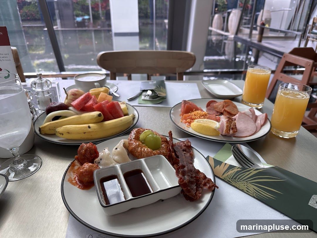 A table laden with a breakfast spread, featuring plates of fresh fruit, assorted meats, bacon, pastries, and glasses of orange juice. A cup of coffee or tea is also visible. The setting appears to be indoors with a view of water and greenery through large windows.