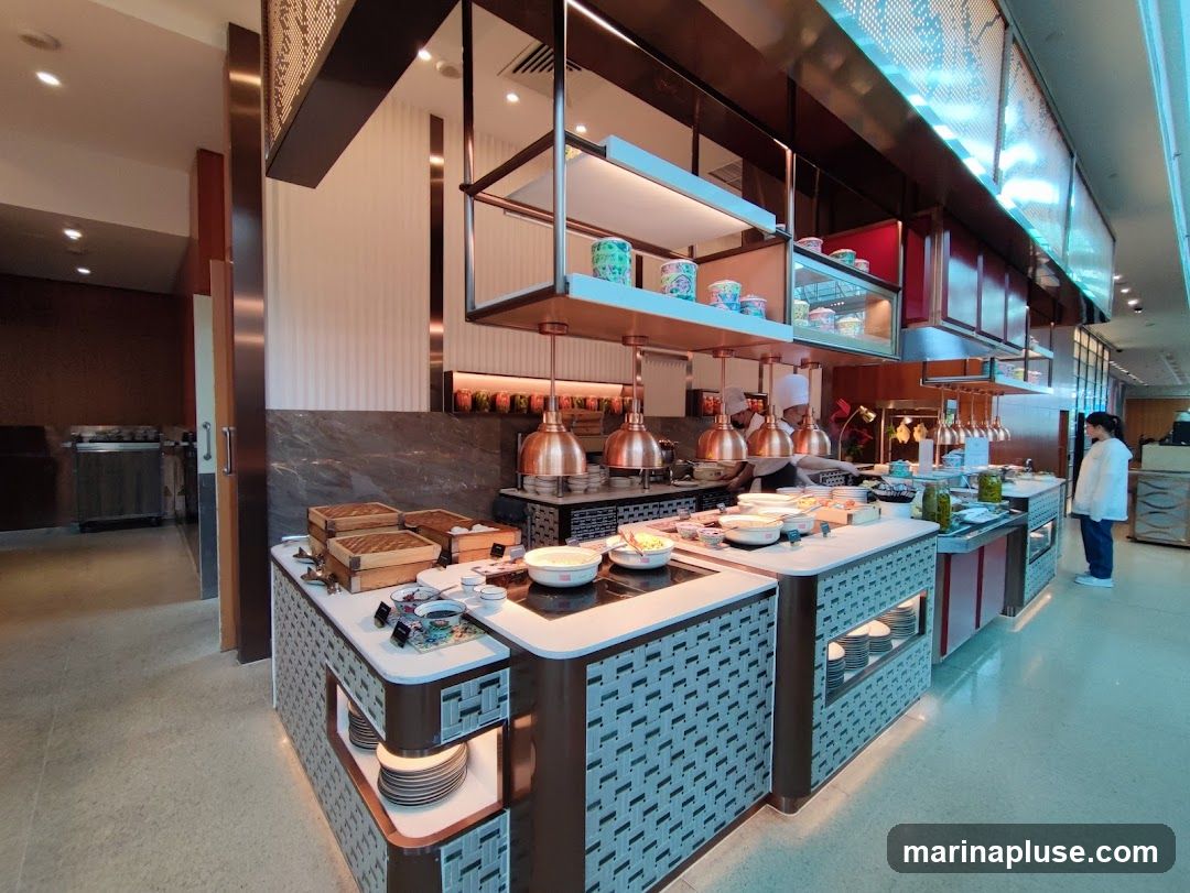 A well-lit buffet station with various food items and serving dishes. Chefs in uniform are visible behind the counter, preparing food. A customer is standing at the end of the buffet, looking at the offerings. The decor is modern with metallic accents and patterned surfaces.