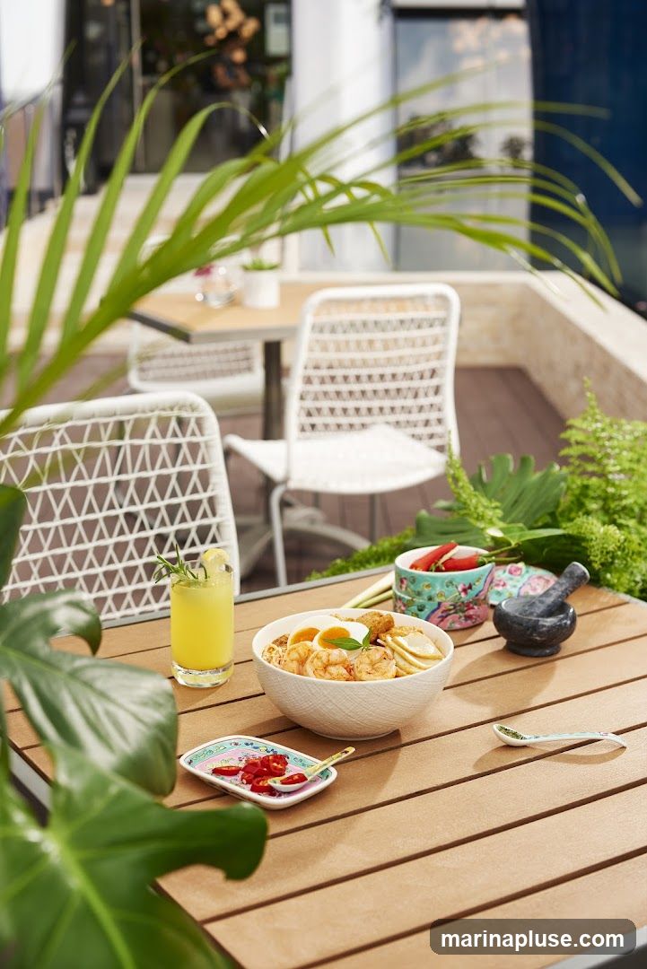A wooden table is set for dining outdoors, with a bowl of noodles, a glass of yellow drink, and a small dish of chili peppers. White chairs and tables are visible in the background, along with lush greenery.