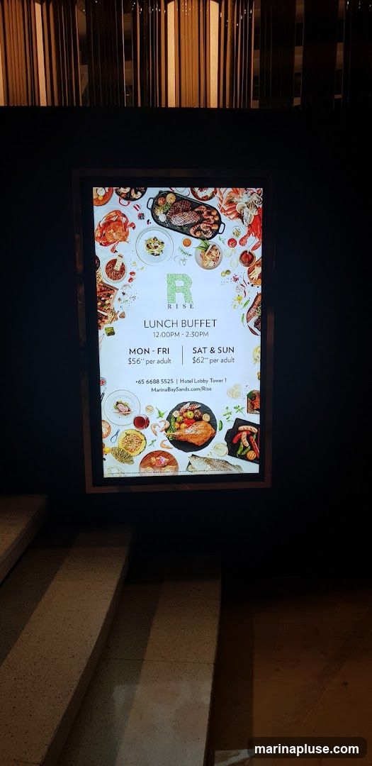 A digital menu board displays information about a lunch buffet. The buffet is available from 12:00 PM to 2:30 PM. The price for Monday to Friday is $56 per adult, and for Saturday and Sunday, it is $62 per adult. Contact information and a website are also provided.