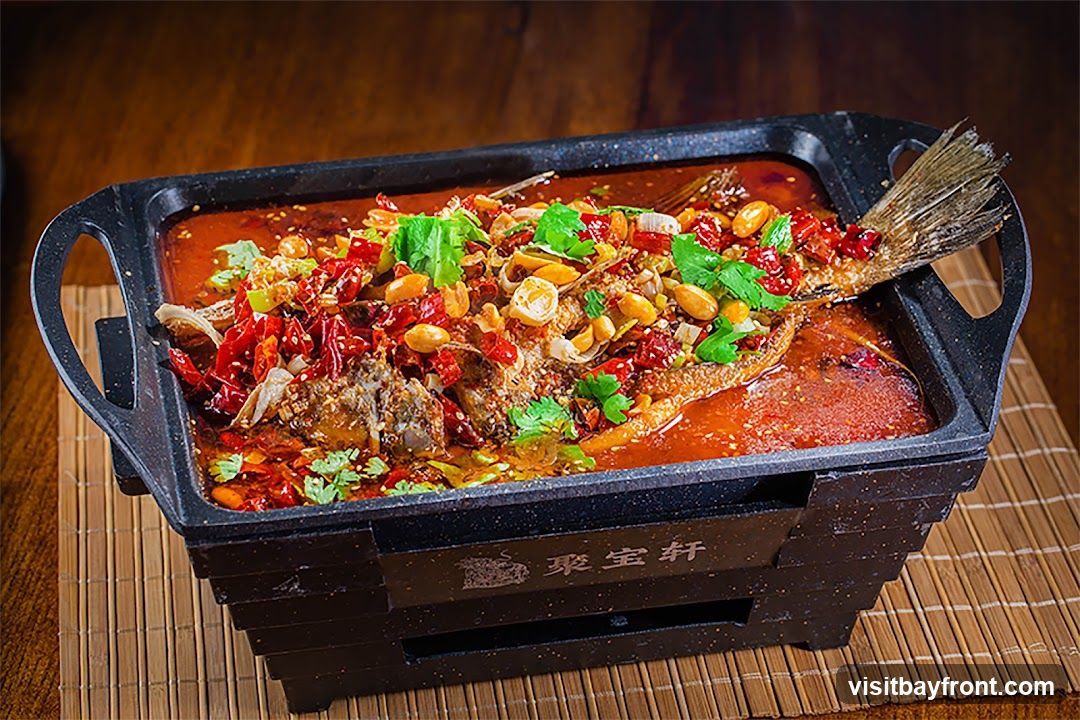 A whole fish is braised in a spicy, red broth, topped with dried chilies, peanuts, and fresh cilantro. The dish is served in a black, rectangular metal pan with handles, which sits on a tiered black stand. The stand has Chinese characters inscribed on it. The pan is placed on a bamboo mat on a wooden surface.