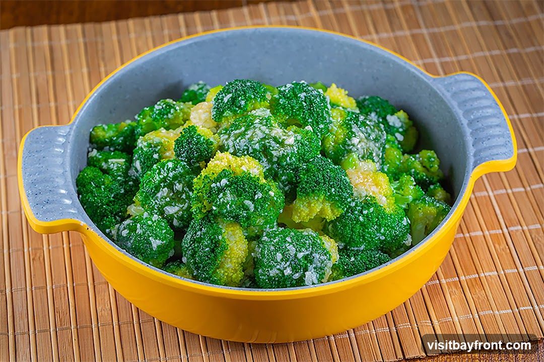 A close-up shot of a yellow and gray bowl filled with cooked broccoli florets. The broccoli appears to be seasoned with salt and possibly garlic. The bowl is placed on a bamboo placemat.