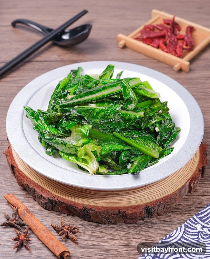 A plate of stir-fried green vegetables, likely a leafy green like bok choy or gai lan, is presented on a wooden table. The vegetables appear tender and glistening, mixed with visible pieces of minced garlic. A white plate with a silver rim holds the dish, which is elevated on a round wooden slice. In the background, a pair of black chopsticks rests on a black spoon, and a small bamboo dish contains dried red chili peppers. Cinnamon stick and star anise are placed on the wooden surface in the foreground, along with a blue and white patterned cloth.