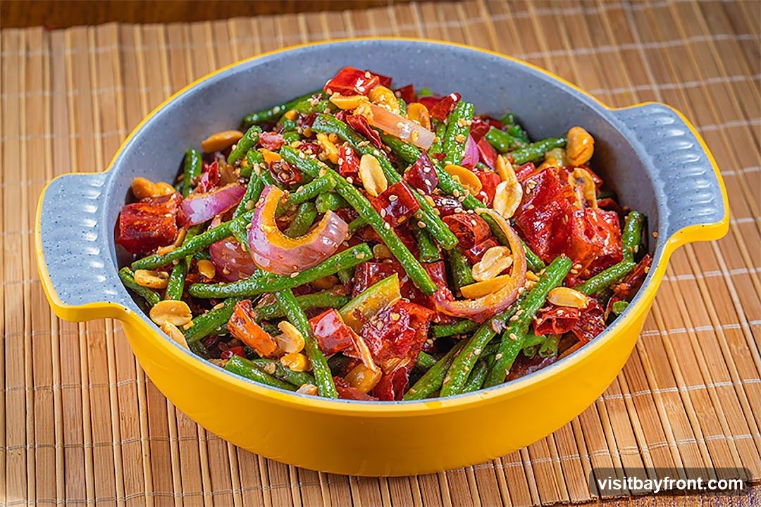 A close-up shot of a yellow and gray bowl filled with stir-fried green beans. The dish is mixed with red chili peppers, red onions, and peanuts, and sprinkled with sesame seeds. The bowl is placed on a bamboo mat.