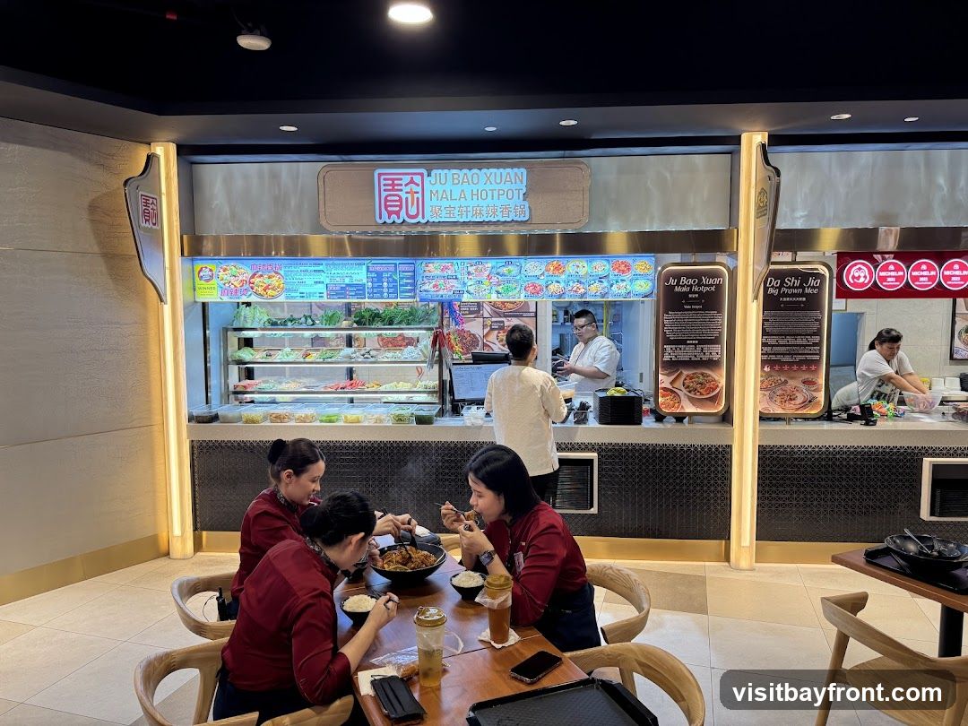 A food stall in a food court with a display of fresh ingredients and a menu board. Several people are seated at a table eating.