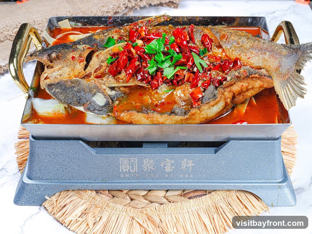 A whole fish, braised in a spicy red sauce and topped with dried chilies and chopped cilantro, is served in a metal tray with gold handles. The tray rests on a grey heating base with Chinese characters and a woven placemat.