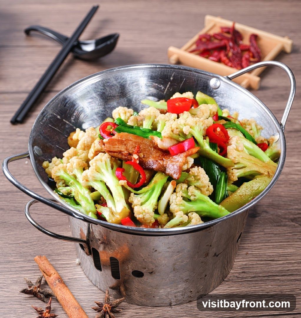 A close-up shot of a metal bowl filled with stir-fried cauliflower, meat, and red chili peppers, served on a wooden table. Chopsticks and a spoon rest in the background, along with a small dish of dried chilies. Cinnamon sticks and star anise are scattered on the table.