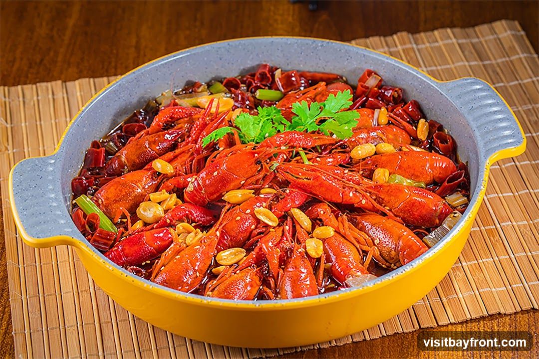 A close-up shot of a large serving of spicy crayfish in a yellow and gray pot. The crayfish are bright red and mixed with dried chili peppers, peanuts, and green herbs. The dish is placed on a bamboo mat.
