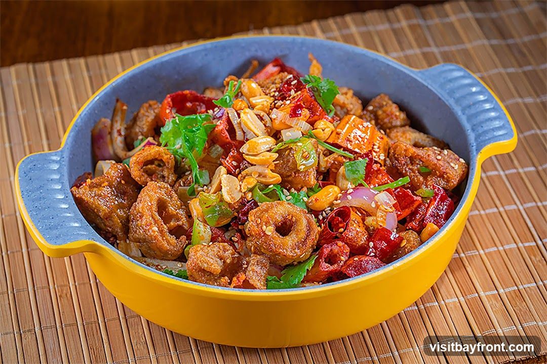A close-up shot of a spicy fried dish served in a yellow and gray bowl. The dish contains pieces of fried meat, red chili peppers, onions, peanuts, and cilantro, all coated in a flavorful sauce. The bowl is placed on a bamboo placemat.