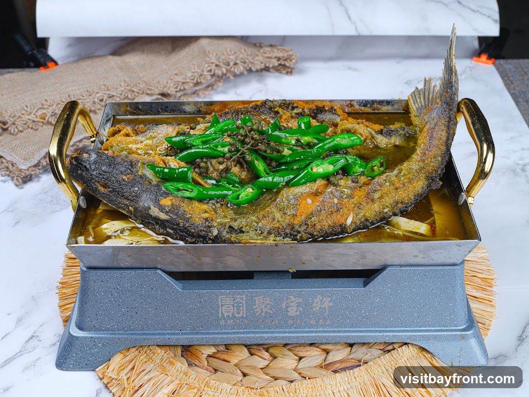 A whole fried fish is served in a rectangular metal dish with golden handles, placed on a grey heating base. The fish is topped with sliced green chilies and other spices. The dish is presented on a woven placemat against a marble-patterned background.