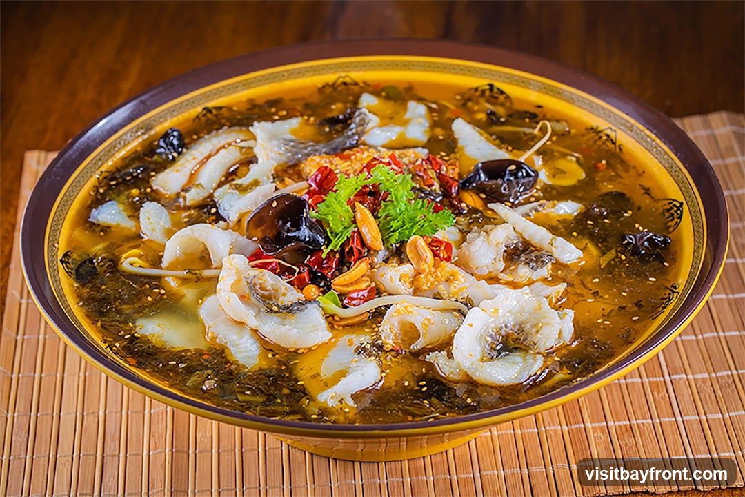 A close-up shot of a large bowl of spicy fish soup. The soup is a rich, oily broth filled with chunks of white fish, pickled vegetables, dried chilies, mushrooms, and peanuts. A sprig of fresh green herbs garnishes the center of the dish. The bowl is placed on a bamboo mat.