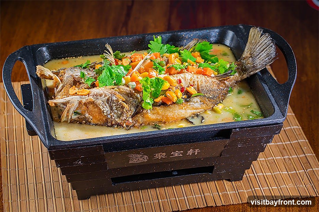 A whole steamed fish is served in a black rectangular dish with handles, placed on a bamboo mat. The fish is topped with chopped carrots, peas, and cilantro. The dish has Chinese characters on the front.