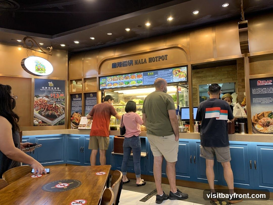 People are queuing at a food counter in a mall. The counter has a sign that says "MALA HOTPOT". There are various food displays and menus visible. Social distancing markers are on the tables in the foreground.