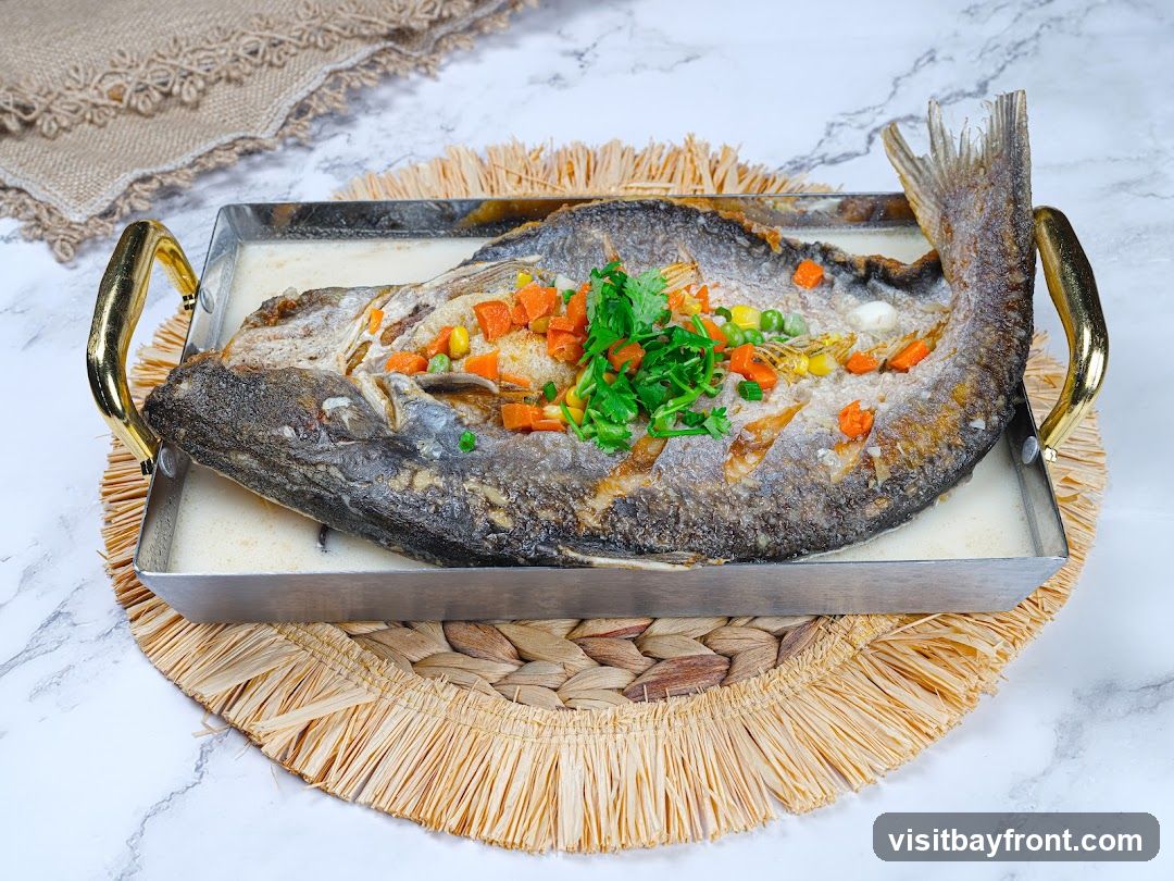 A whole steamed fish is presented in a rectangular metal dish with gold handles, placed on a woven placemat. The fish is topped with chopped carrots, peas, and fresh cilantro. The dish is set against a marble-patterned background with a textured fabric runner in the upper left corner.